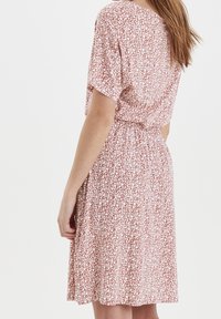 Květinové šaty v cihlově červené barvě s bílými listovými vzory. Krátké rukávy, elastický pas a délka ke kolenům. Lehká látka s volným střihem.