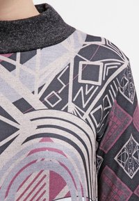 Haut coloré à motifs avec un col gris ; présente des designs géométriques en rose, noir et beige. Tissu doux et texturé avec des détails complexes.