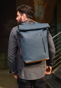 Rucksack mit Rolltop aus dunkelblauem wasserdichtem Material, mit einem braunen Lederakzent an der Basis und einer mit Reißverschluss versehenen Vordertasche.