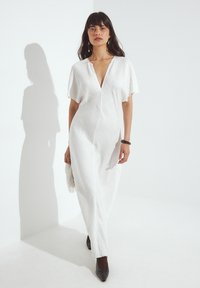Bela obleka z V-izrezom, kratkimi rokavi in teksturirano površino. V kombinaciji s temnimi špicastimi čevlji in torbico z resicami. Preprosta, elegantna zasnova.