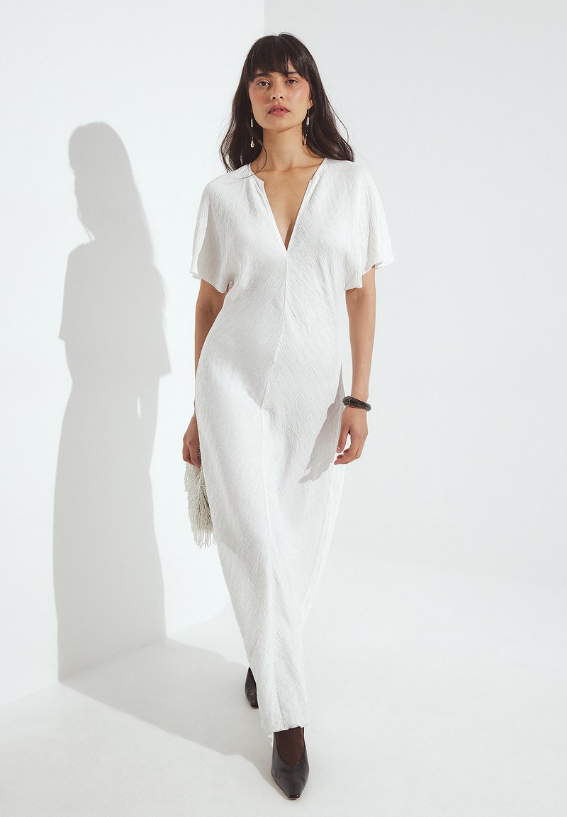 Bela obleka z V-izrezom, kratkimi rokavi in teksturirano površino. V kombinaciji s temnimi špicastimi čevlji in torbico z resicami. Preprosta, elegantna zasnova.