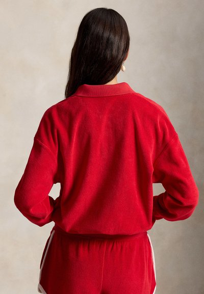 Femme aux longs cheveux foncés vue de dos, portant un haut rouge à manches longues et un short rouge assorti avec des bordures blanches.