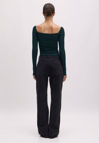 Langärmliges, dunkelgrünes Oberteil mit quadratischem Ausschnitt, kombiniert mit hochgeschnittenen, schwarzen weit geschnittenen Jeans. Glatte Textur, figurbetonte Silhouette.