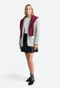 Blazer gri din lână, suprapus cu un hanorac cărămiziu, asortat cu o fustă plisată neagră și loafers negri. Un top alb și șosete gri completează ținuta.