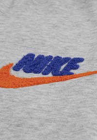 Pelēka auduma ar teksturētu, izšūtu "NIKE" logo zilā krāsā un oranžā swosh. Abiem logotipiem ir kontrastējošas dzīvīgas krāsas un dziļums.