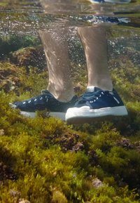 Jambes portant des baskets bleu marine, debout sous l'eau parmi des algues vertes et brunes avec des bulles d'air qui montent.