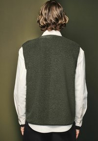 Dark green, textured vest with a round neckline and no sleeves, layered over a white long-sleeve shirt. Smooth fabric with a relaxed fit.