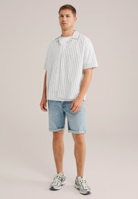 Moški stoji oblečen v kratko majico z belimi črtami čez belo majico, v svetlobluščenih zavihanih jeans kratkih hlačah in belih supergah.