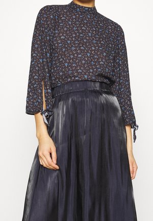Kobieta w czarnej kwiecistej bluzce z niebieskimi kwiatami i wiązanymi rozcięciami na rękawach, w połączeniu z wysokim stanem, błyszczącą czarną plisowaną spódnicą.