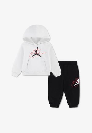 Biała bluza z kapturem z czarnym i czerwonym logo Air Jordan, w połączeniu z czarnymi dresowymi spodenkami z elastycznym pasem i pasującym logo na nodze.