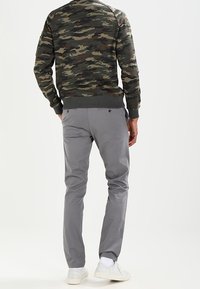 Felpa con motivo camouflage in verde scuro e marrone, abbinata a pantaloni grigi chiari e sneakers bianche, vista da dietro.