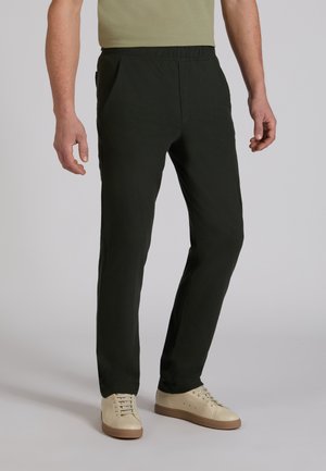 Uomo che indossa pantaloni slim fit verde scuro, sneaker beige e una t-shirt verde oliva su sfondo grigio chiaro uniforme.