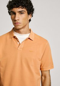 Oranžs polo krekls izgatavots no kokvilnas ar fakturētu apdari. Iezīmē klasisku apkakli, divus pogas un izšūtu logo kreisajā pusē.