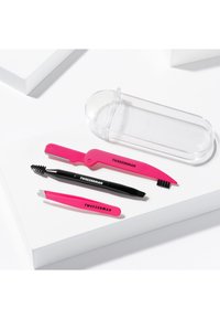 Trois outils de soin des sourcils Tweezerman roses et noirs avec un étui en plastique transparent sur une surface blanche.