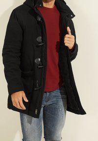Abrigo negro con capucha, con cierre de botones, dos bolsillos laterales y un acabado texturizado, superpuesto a una camisa roja y unos vaqueros azules.