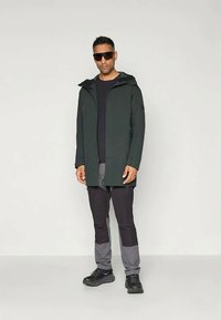 Σκούρο πράσινο αδιάβροχο μπουφάν με κουκούλα, συνδυασμένο με μαύρο και γκρι παντελόνι. Διαθέτει πλαϊνές τσέπες και κομψό, σύγχρονο σχέδιο. Μαύρα παπούτσια.