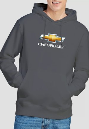 A man smiles wearing a gray hoodie featuring a large Chevrolet logo across his chest, paired with blue jeans against a neutral background.