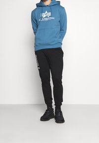 Blå hoodie med vit logotyp, kenguruficka och justerbar huva; kombinerad med svarta smalrandiga sweatpants med vit text; svarta sneakers.
