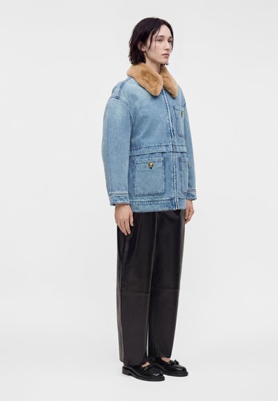 Personne portant une veste en jean bleu clair avec un col en fourrure marron, un pantalon noir à jambes larges et des mocassins noirs, se tenant devant un fond uni.