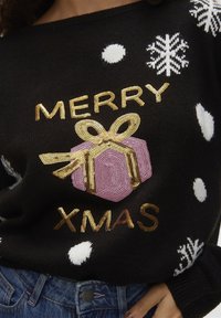 Svart stickad tröja med texten "MERRY XMAS" i guldpaljetter, en rosa presentförpackning med guldaccenter och mönster av vita snöflingor och prickar.