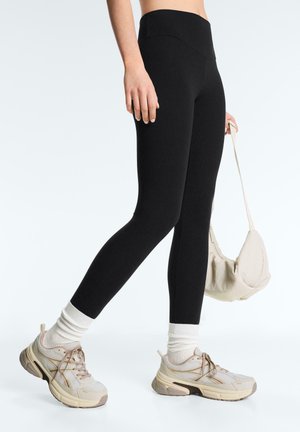 Person trägt schwarze Leggings, weiße Socken, beigefarbene Sneakers, hält eine kleine beigefarbene Umhängetasche und steht vor weißem Hintergrund.