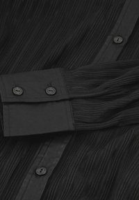 Black pleated shirt with long sleeves, featuring textured fabric, black buttons, and a classic collar, emphasizing a wrinkled pattern.