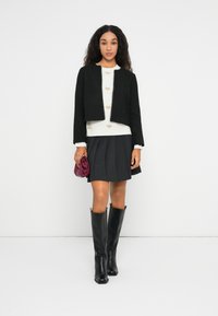 Kvind med langt, krøllet hår iført sort cropped jakke, hvid sweater med hjerte mønstre, plisseret sort nederdel, sorte knæhøje støvler, holder en lilla clutch.