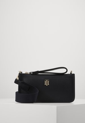 Black rectangular leather wristlet with gold "TH" logo and detachable wide fabric shoulder strap on white surface.