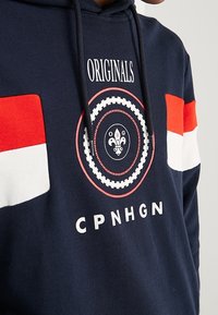 Felpa navy con accenti rossi e bianchi, caratterizzata da un logo circolare con un design floreale e il testo "ORIGINALS" e "CPNHGN."