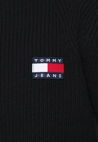 Suéter de punto acanalado negro con un parche rectangular de logo que lleva "TOMMY" y "JEANS" en letras blancas y rojas sobre un fondo marino.
