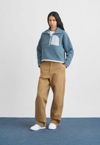 Μπλε φούτερ με μισό φερμουάρ, τσέπη στο στήθος και λευκή λεπτομέρεια; συνδυασμένο με καμπάνα παντελόνι χακί και λευκά αθλητικά παπούτσια σε μπλε φόντο.