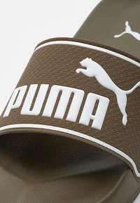 Bruna gummiskosandaler med en texturerad ovansida, vit "PUMA"-logotyp och en hoppande katt-emblem. Mjuk sula och öppen tå-design.