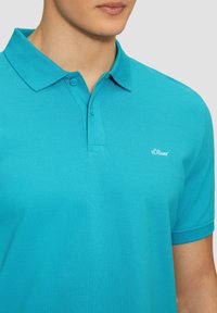 Türkise polo-särk, mis on valmistatud tekstureeritud kangast, sisaldab kahe nupuga kaelust ja väikest valget logo vasakul rinnal.