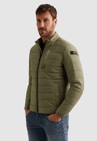 Hombre con barba vistiendo una chaqueta acolchada verde claro con parches en el pecho y la manga, con las manos en los bolsillos, de pie frente a un fondo liso.