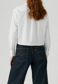 Chemise blanche courte à manches longues, associée à un pantalon en denim bleu foncé avec des poches arrière et des détails de surpiqûres jaunes.