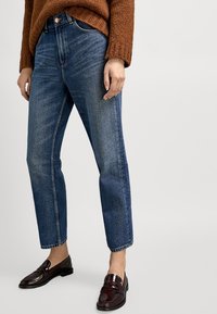 Jeans in denim blu con gamba affusolata, vita alta e texture sbiadita; abbinati a mocassini marrone scuro e un maglione in maglia marrone.