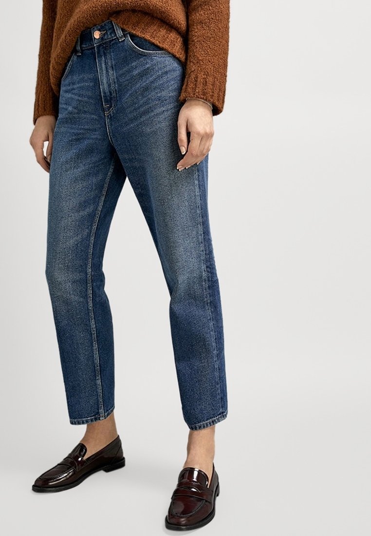 Jeans in denim blu con gamba affusolata, vita alta e texture sbiadita; abbinati a mocassini marrone scuro e un maglione in maglia marrone.