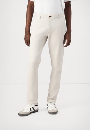 Beige cotton trousers with a slim fit, featuring a button closure and pockets. Paired with gray and white sneakers with black accents.