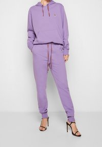 Fialová mikina s klokaní kapsou, nastavovací šňůrkou a vyšitým detailem zvířete. V kombinaci s odpovídajícími tepláky, oboje vyrobené z měkké látky.