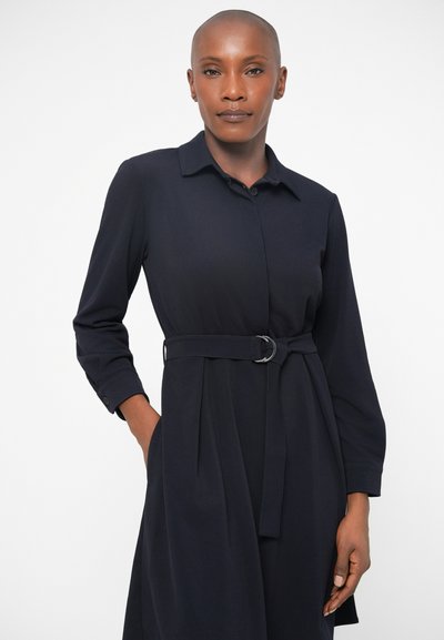 Moteris su nuskustais plaukais, vilkinti ilgomis rankovėmis tamsiai mėlynos spalvos suknelę su apykakle ir diržu, stovi su viena ranka kišenėje priešais baltą foną.