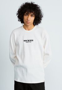 Biała prążkowana koszulka z długim rękawem i okrągłym dekoltem. Na przodzie znajduje się czarny napis "DICKIES since '22". Miękka w dotyku, luźny krój.