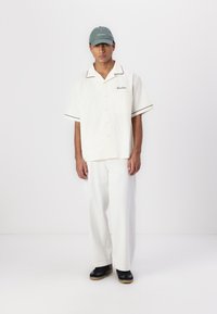 Short-sleeve cream shirt with black trim, paired with loose white pants and black shoes. The cap is green with a logo.