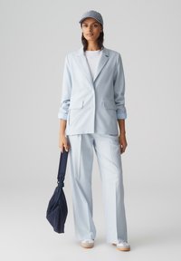 Blazer azzurro chiaro con due tasche frontali, abbinato a pantaloni coordinati. L'outfit è completato da una t-shirt bianca, un cappellino a righe e sneakers.
