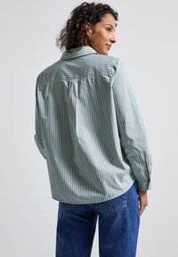Vrouw in een lichtgroen en wit gestreept shirt met lange mouwen en blauwe jeans, iets naar opzij gekeerd tegen een effen achtergrond.