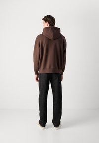 Brown hoodie with a large hood, paired with dark grey pants and light-colored shoes. The fabric appears smooth with a relaxed fit.