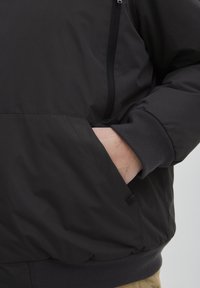 Jachetă neagră, izolată, cu front cu fermoar, manșete ribbate și buzunare laterale. Materialul pare neted, cu un finisaj mat.