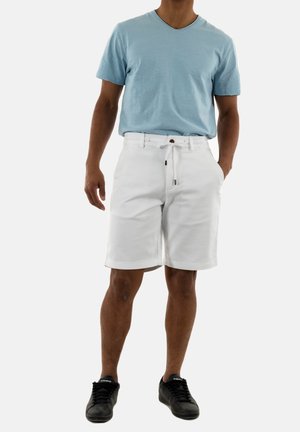 Pantalones cortos de algodón blancos con bolsillos, cintura elástica y cordón ajustable. Combinados con una camiseta de manga corta y corte en V azul y zapatos negros.