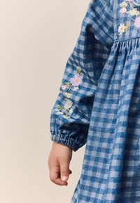 Mėlyna languota suknelė su pūstais rankovais, siuvinėtais gėlių akcentais ant rankogalių ir lygiu audiniu, vidutinio ilgio dizainu.