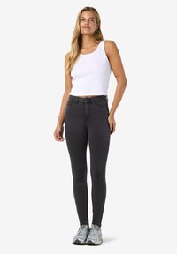 Juodos spalvos siauri džinsai su aukštu liemeniu, derinami su balta ribuota liemene ir pilkais sportiniais batais, pabrėžiantys prigludusį dizainą ir lygią tekstūrą.