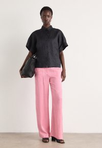 Camicia nera in lino a maniche corte con bottoni abbinata a pantaloni rosa chiaro a gamba ampia. Tiene una borsa nera con trama. Design minimalista.